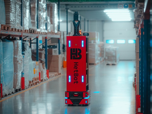 Autonomous HetBot mobile robot navigating independently in a warehouse between shelves with goods.